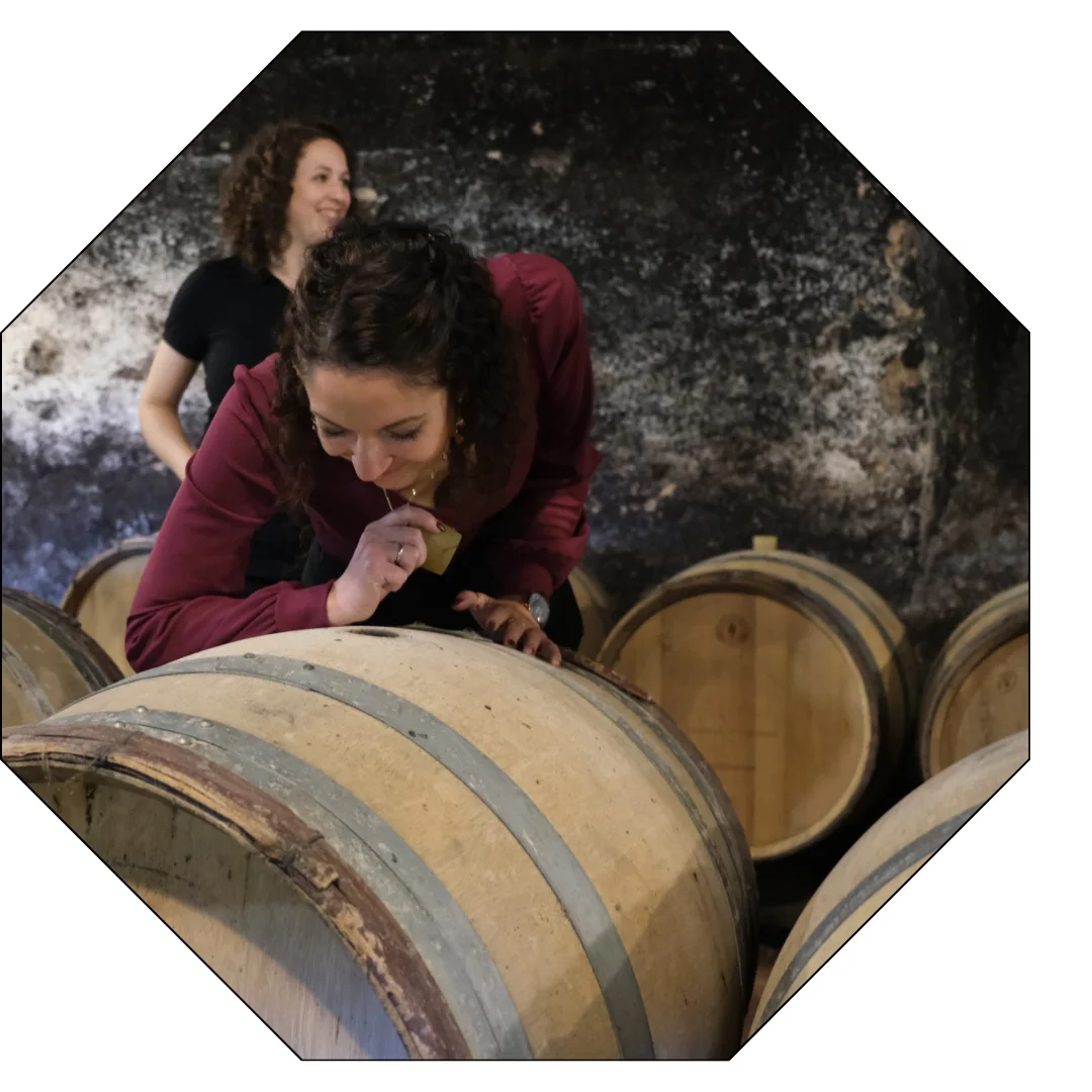 Pendant qu'Inès Delmotte présente la cave, une jeune femme découvre les arômes du vin qui se dégagent d'un tonneau lors d'une visite immersive dans un château du Beaujolais.
